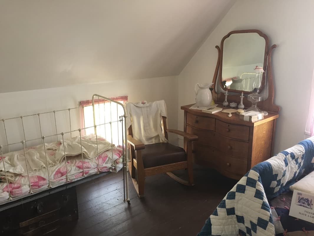 An upstairs bedroom under a sloped ceiling with an iron crib, a christening gown on a rocker and quilts