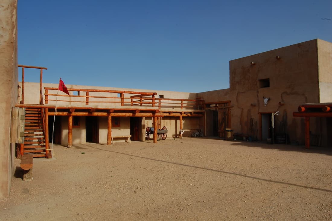 The fort courtyard in full sun: a two-level log balcony and staircase, a red flag, a wagon and a bison skull on an adobe tower