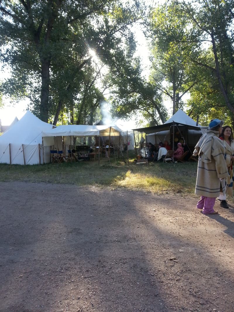 White wall tents under backlit cottonwoods with campfire smoke, and women in blanket capotes