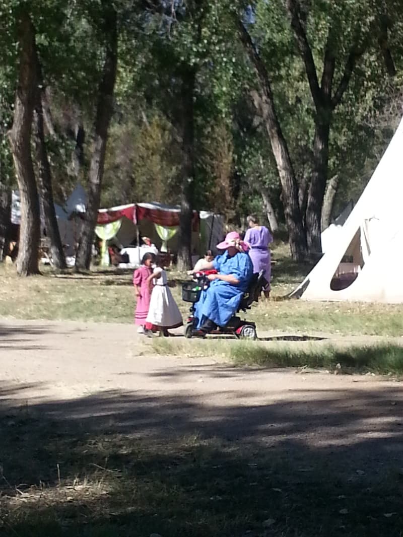 A woman in a blue dress with two girls in period dresses beside a tipi