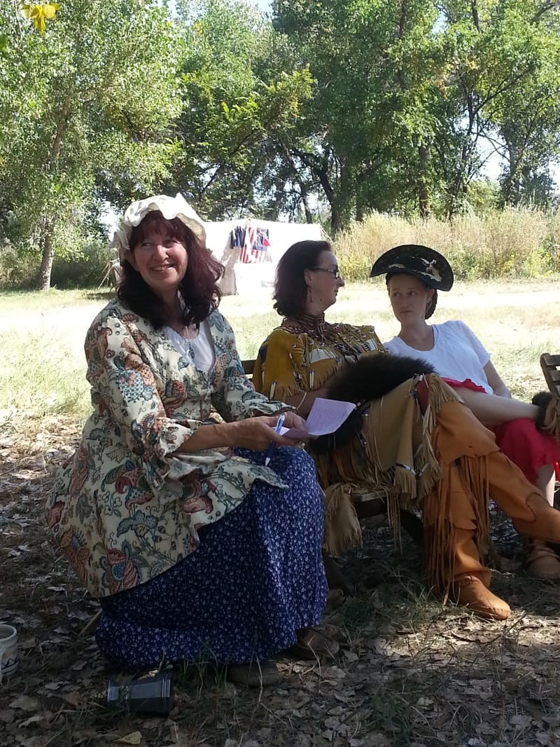 Three women in period dress, one in a calico jacket and mob cap, seated on a bench and smiling