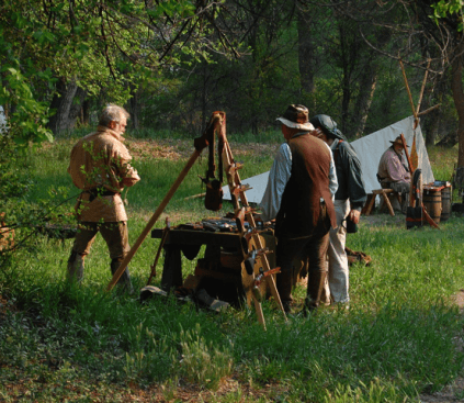 Reenactors in period clothing gathered at a trade table in a spring-green meadow