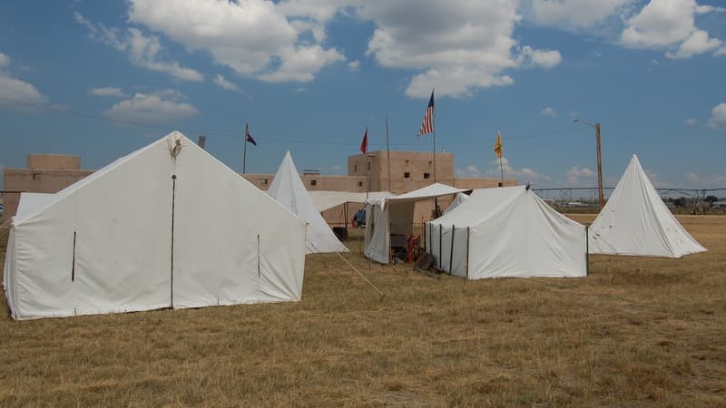A rendezvous camp of white wall tents and tipis on dry grass with flags flying and the fort behind