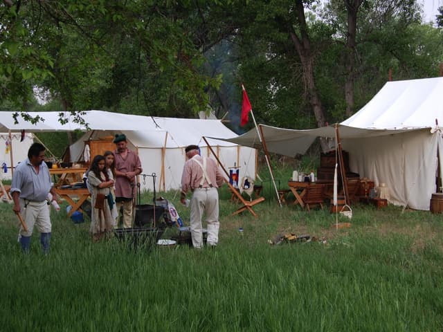 Reenactors and children in period dress around a cook fire and kettles, with wall tents and flags