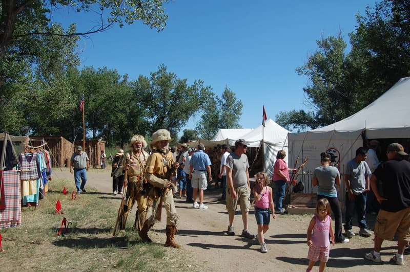 Two mountain men in buckskins and fur hats walking a busy rendezvous lane among families and trader tents