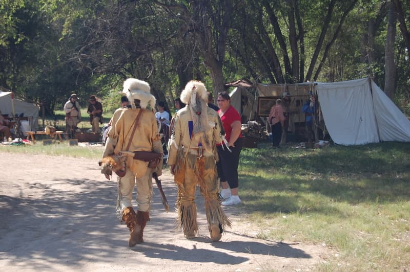 Two mountain men seen from behind in fringed buckskins and wolf-fur hats, walking toward the trader tents