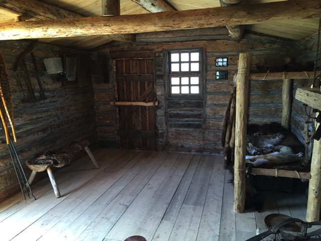 Inside the cabin: a plank door, a small window and a fur-covered bunk and stool on a plank floor