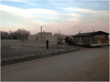 The trapper cabin on a flatbed trailer arriving at the park at dusk, with people watching and the fort behind