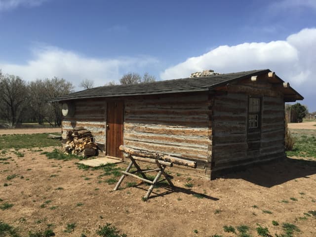 The restored 1850s trapper cabin at the park, hewn logs with white chinking, a woodpile and a sawhorse under a big sky